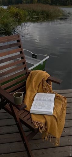 an open book is sitting on a wooden chair by the water with a yellow blanket draped over it