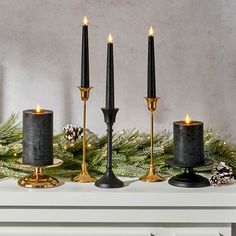 three black candles sitting on top of a mantle next to evergreen branches and pine cones