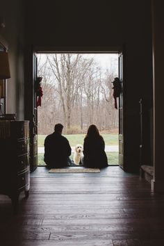 two people sitting on the floor in front of an open door looking at a dog