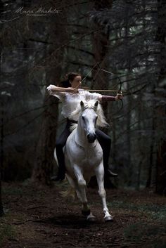 a woman riding on the back of a white horse through a forest filled with trees