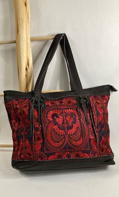 Pranee Phoenix Collection Sierra in Black      #100-G Sierra, from the Phoenix Collection, is a soft black leather tote with hand-sewn embroidery (front to back) in a bold mythical design paired with a bold color palette: red, blue, and purple to name a few. Pranee Handcrafted Unique Designer Bags are beautiful artisan-made bags completely handmade using genuine leather, hand sewn embroidery, and unique decorative stitching.  There is an incredible amount of care and attention to details taken in the craftsmanship of every Pranee bag.  Each piece is made with genuine leather, hemp or cotton, and unique hand-sewn embroidery and/or applique. Features *        Zipper closure *        Interior side pockets and zipper pocket *        Genuine leather handle and accents  Specifications *        D Traditional Black Tote Shoulder Bag, Artisan Black Tote Bag, Hand-stitched Travel Bags, Artisan Black Bag With Leather Handles, Artisan Black Bags With Leather Handles, Leather Hand-stitched Shoulder Bag, Black Hand Tooled Tote Bag, Black Hand Tooled Bags For Everyday Use, Artisan Black Tote Shoulder Bag