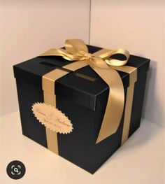 a black box with gold ribbon and a bow on it's side, sitting on a white surface