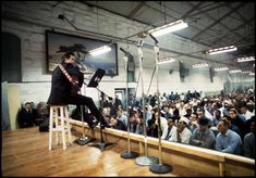 a man sitting on top of a stool in front of a microphone with an audience behind him