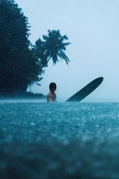 a person standing in the water with a surfboard under their arm, and trees behind them