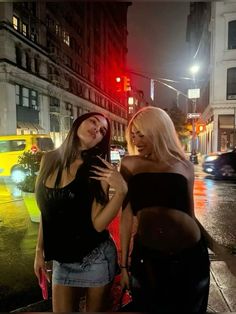 two beautiful women standing next to each other on a city street in the rain at night