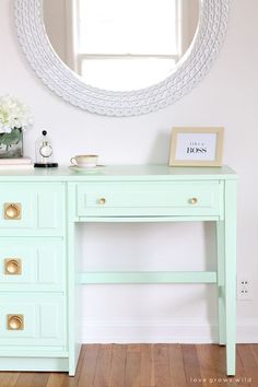 a mint green desk with drawers and a mirror on the wall in front of it