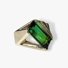 A one-of-a-kind vintage cocktail ring boasting a vibrant elongated emerald cut green tourmaline weighing 16.77 carats set on an angle while accented by 5 round diamonds weighing 0.32 carats set in solid 14k yellow gold with tiered detailing. Complimentary resizing is available up to 3 sizes larger or smaller than the stated size. WEB ID: R603 Luxury Green Tourmaline Emerald Ring, Unique Green Emerald Ring For Formal Occasions, Modern Formal Green Emerald Ring, Modern Green Emerald Ring For Formal Occasions, Modern Green Jewelry For Formal Occasions, Modernist Green Ring For Formal Occasions, Modern Green Tourmaline Jewelry, Vintage Cocktail Ring, Vintage Cocktail