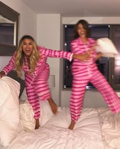 two women in matching pink and white pajamas jumping on a bed while one holds a pillow