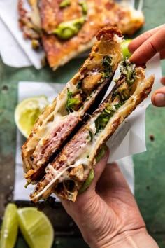 a hand holding a sandwich with meat, cheese and vegetables next to sliced limes