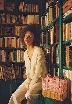 a woman sitting in front of a bookshelf holding a pink bag and looking at the camera