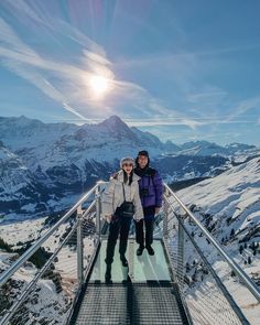 two people standing on a glass walkway in the snow with mountains in the background at sunset