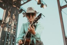 a woman in a hat and sunglasses playing guitar at an outdoor music festival with lights behind her