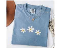 This is a Comfort Colors garment-dyed embroidered daisies t-shirt; a spring tee made 100% with ring-spun cotton. The soft-washed, garment-dyed fabric brings extra coziness to your wardrobe while the relaxed fit makes it an excellent daily choice. The double-needle stitching throughout the embroidered flower tee makes it highly durable while the lack of side-seams helps this cute gift for her shirt retain its tubular shape. .: 100% ring-spun cotton .: Medium fabric (6.1 oz/yd² (206.8 g/m .: Relaxed fit .: Sewn-in twill label PRINT - This is an embroidered daisy shirt, meaning the design is sewn into the fabric.  SIZING - For oversized look, size up 2! WHEN CAN I EXPECT MY ORDER - Processing Time: 1-3 days - During holidays please expect delays as the number of orders are slightly higher tha Blue Tops With Custom Embroidery For Spring, Cotton Daisy Print Tops For Spring, Custom Embroidered Cotton Tops For Spring, Machine Embroidered Short Sleeve Top For Spring, Spring Cotton Top With Custom Embroidery, Short Sleeve Tops With Machine Embroidery For Spring, Spring Custom Embroidered Relaxed Fit Top, Spring Relaxed Fit Top With Custom Embroidery, Spring Cotton Tops With Embroidered Graphics