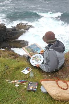 a man sitting on top of a grass covered hillside next to the ocean holding a painting