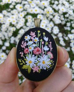 a hand holding a small embroidered pendant with daisies and flowers on it in front of some daisies