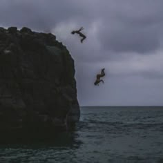 two people jumping off the rocks into the ocean on a cloudy day with dark clouds