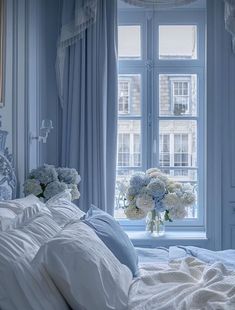 a bedroom with blue walls and white bedding has flowers in a vase on the window sill