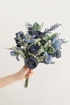 a hand holding a bouquet of blue flowers and greenery on a white wall background