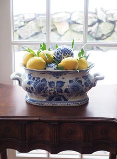 a blue and white bowl filled with lemons on top of a table next to a window