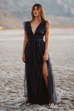 a woman standing in the middle of a desert wearing a long black dress with an open slit