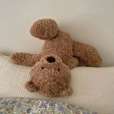 a brown teddy bear laying on top of a bed next to a white wall and pillows