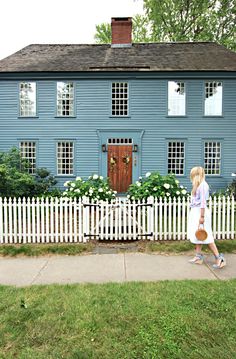 a woman walking down the sidewalk in front of a blue house with white picket fence