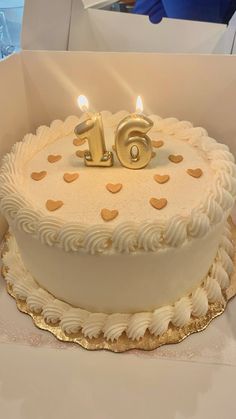 a white cake with gold decorations and two lit candles on top, in a box