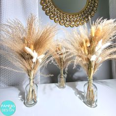 three glass vases with dried plants in them on a white countertop next to a mirror