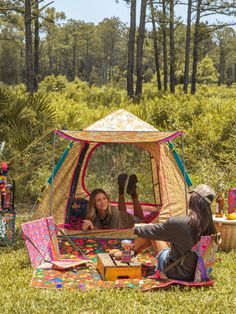 two women sitting in a tent on the ground with food and drinks inside it, surrounded by pine trees