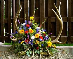 an arrangement of flowers and deer antlers on gravel in front of a wooden fence