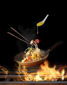 a wok filled with noodles and vegetables being cooked over an open flame on a black background