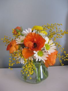 an orange and white flower arrangement in a glass vase on a table with yellow flowers