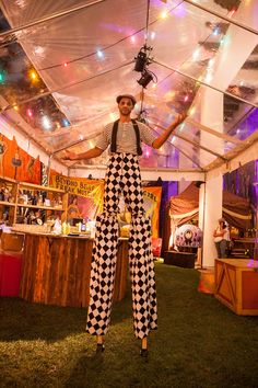 a man in checkered overalls and suspenders stands under a tent with lights