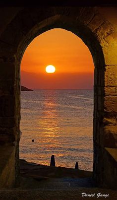 the sun is setting over the ocean through an arch in a stone wall with water below it