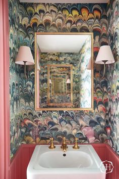 a bathroom sink under a mirror in front of a wallpapered wall and lamp