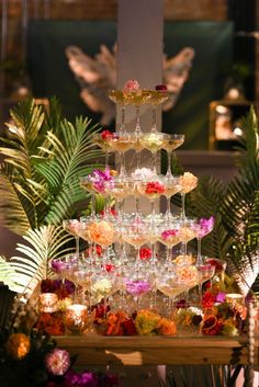 a tall tower filled with lots of wine glasses next to flowers and greenery in front of a statue