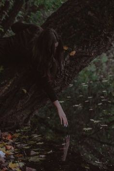 a woman leaning up against a tree in the woods with her reflection on the ground