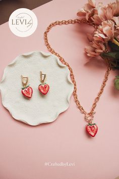 two strawberry shaped earrings and necklace on a pink surface next to a white plate with flowers