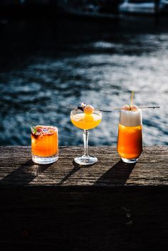 three drinks sitting on top of a wooden table next to each other near the water