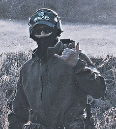 a man wearing a black jacket and hat standing in the grass with his finger up