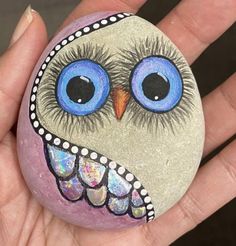 a hand holding a painted rock with an owl's face and blue eyes on it