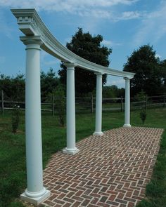 a brick walkway between two white pillars in a grassy area with trees and blue sky