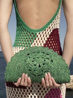 a woman holding a crocheted purse in front of the ocean with her back to the camera