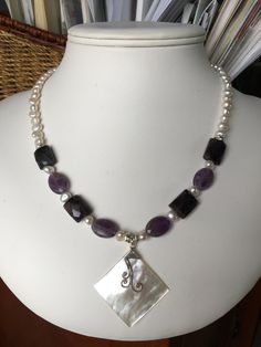 The focal point of this necklace is a rectangular pendant of white Mother of Pearls, which was handcrafted in Bali. The strand combines faceted ovals and faceted of rectangles of amethyst, grey freshwater pearls and silver accents. The necklace is 18 inches long, is strung on jeweler's wire and has a sterling silver lobster clasp. Item number 0519-SN8 White Amethyst Pendant Jewelry, Handmade Purple Rectangular Necklace, Elegant Purple Rectangular Necklace, Rectangular Purple Amethyst Necklaces, Purple Rectangular Amethyst Necklaces, Purple Rectangular Amethyst Necklace, Faceted Silver Pearl Necklace For Gift, Elegant Rectangular Gemstone Beads Jewelry, White Amethyst Pendant Necklace