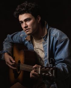 a young man is playing the guitar and looking at the camera while wearing a denim jacket