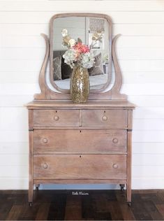 a wooden dresser with a mirror and flowers on it's top, in front of a white wall