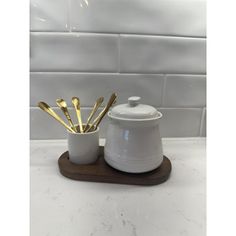 a white ceramic container with gold spoons in it on a wooden tray next to a tiled wall