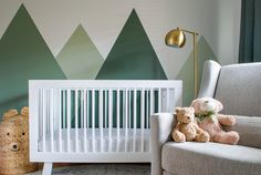 a baby crib with a teddy bear sitting next to it in front of mountains painted on the wall