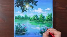 a person is painting a landscape with water and trees on the side of a wooden table