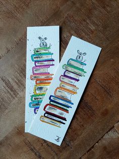 two bookmarks with colorful books on them sitting on a wooden table next to each other